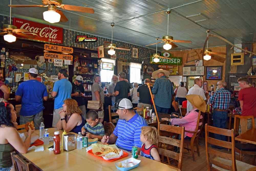 A lively dining area is filled with patrons enjoying meals and drinks. Wooden tables are set with plates and beverages. In the background, a bar is lined with a variety of bottles and vintage signs, contributing to the rustic atmosphere of the space.