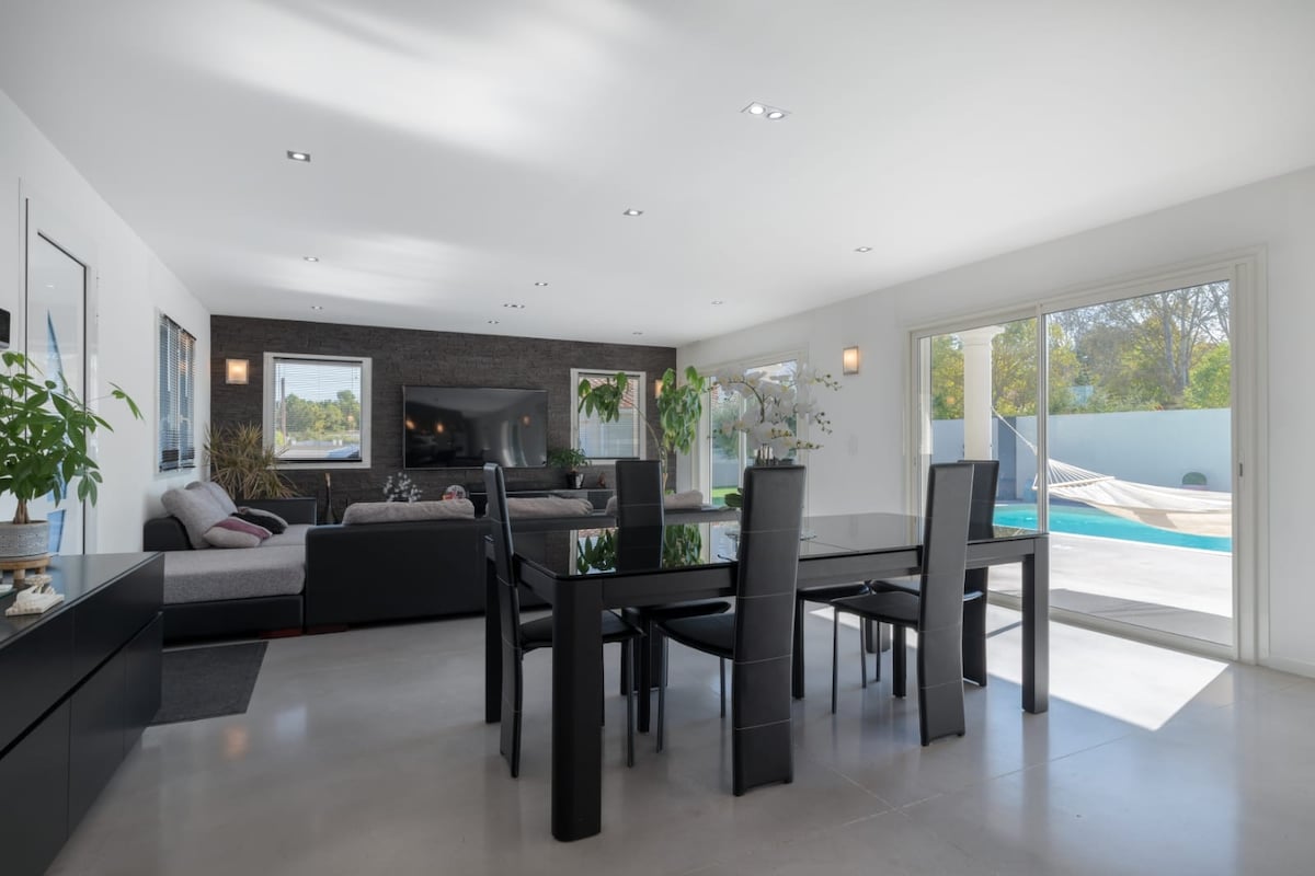 An open living space features a modern dining table surrounded by six sleek chairs. Natural light fills the area from large sliding glass doors, providing a view of the pool and backyard. A comfortable seating area is visible in the background.
