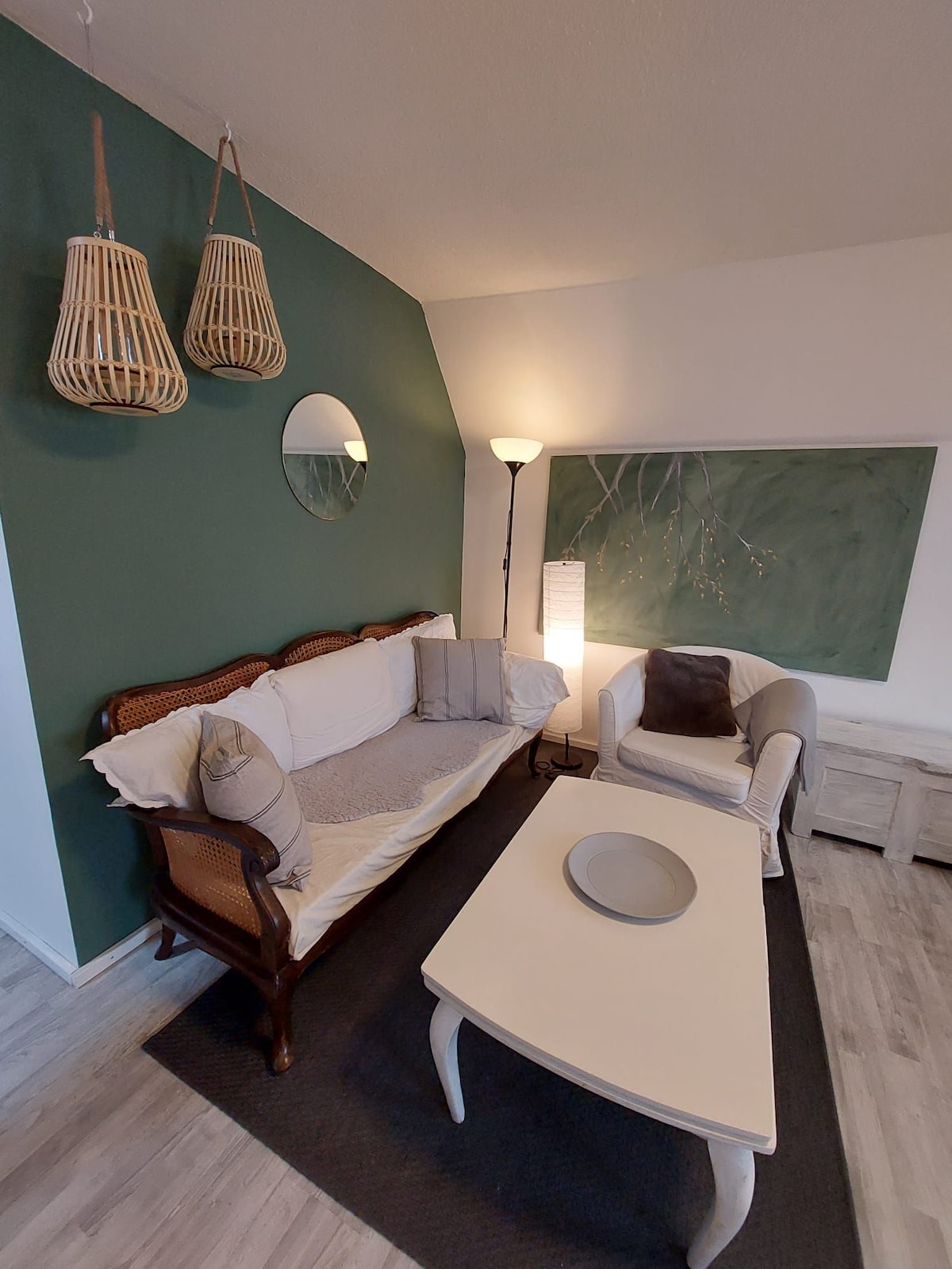 A cozy living area is showcased, featuring a rattan sofa and a light armchair. A round mirror is mounted on a green accent wall, while a lamp provides warm lighting. A light coffee table sits atop a dark area rug, complementing the neutral color palette.