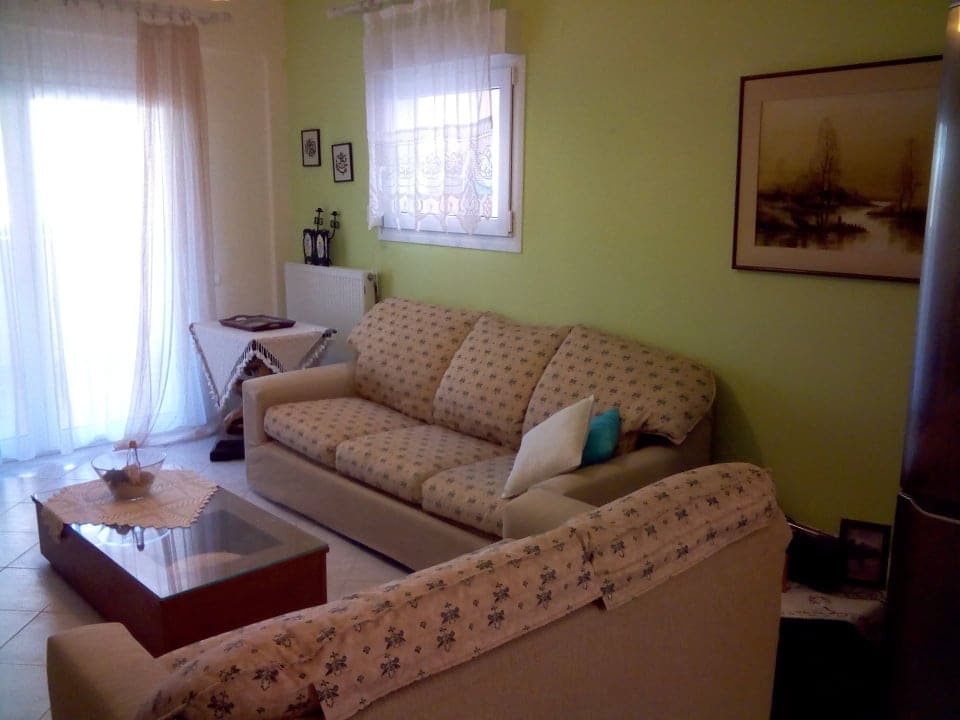 A comfortable living area features a light-colored sofa and armchair, arranged around a wooden coffee table. Natural light enters through glass doors that lead to a small terrace, and a vibrant green wall contrasts with the neutral furnishings. Framed art pieces and decor are displayed on the walls.