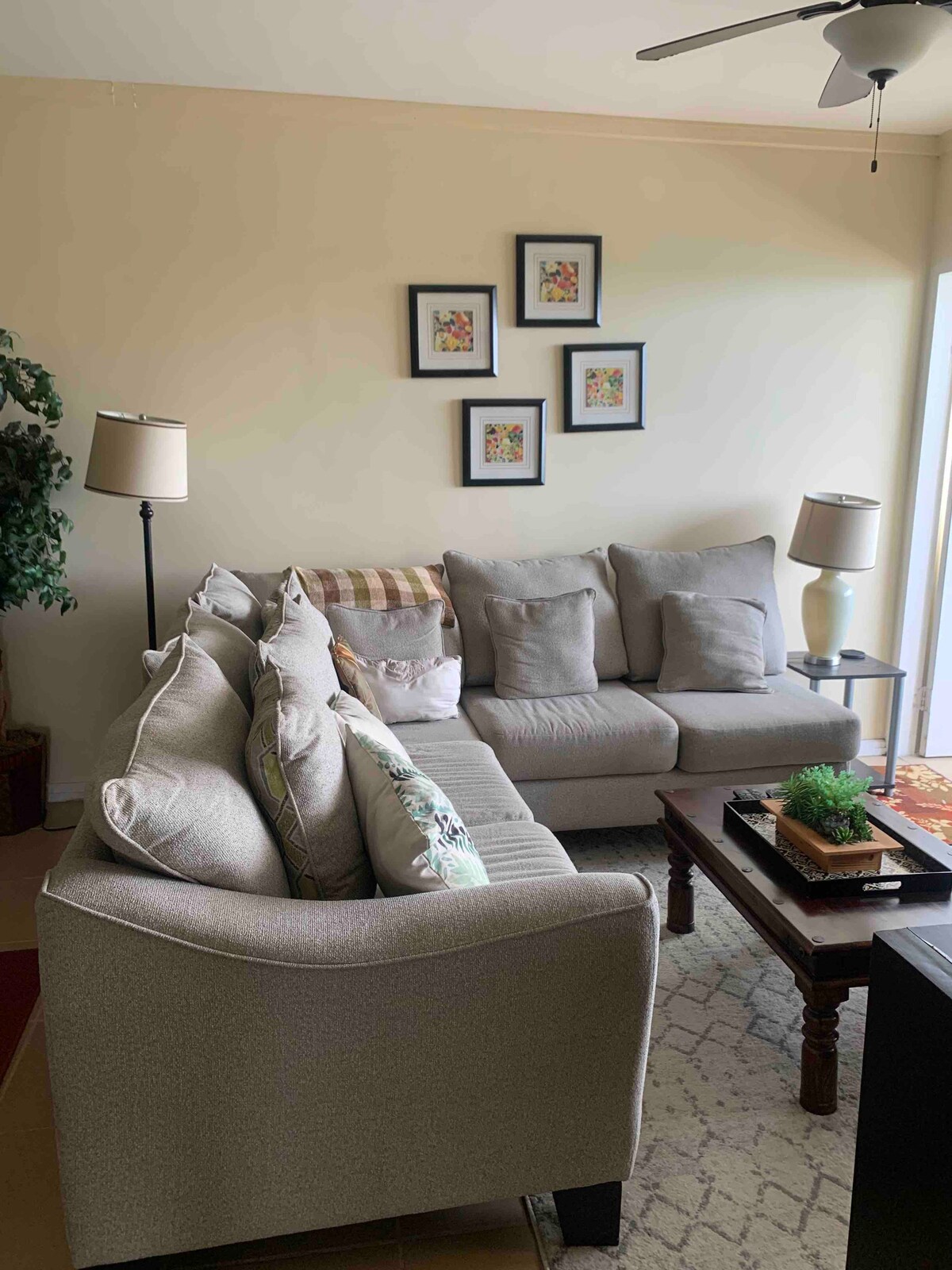 A comfortable living area features a light gray sectional sofa adorned with decorative pillows. A coffee table sits in front, showcasing a small plant and decorative items. Two floor lamps are positioned nearby, and framed artwork decorates the wall above the sofa.