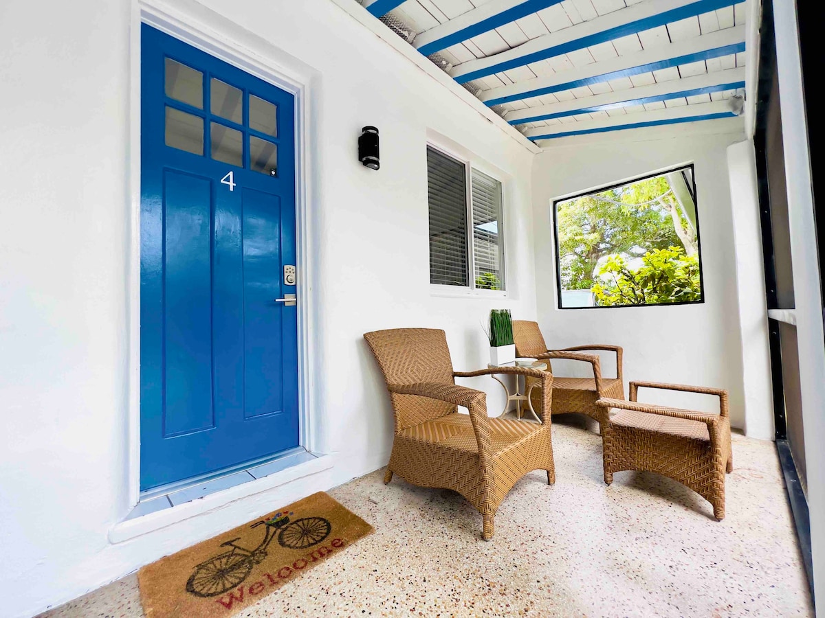 A private entrance is showcased, featuring a vibrant blue door and two wicker chairs positioned around a small table. A welcome mat is placed at the doorway. Natural light filters through a large window, revealing lush greenery outside.
