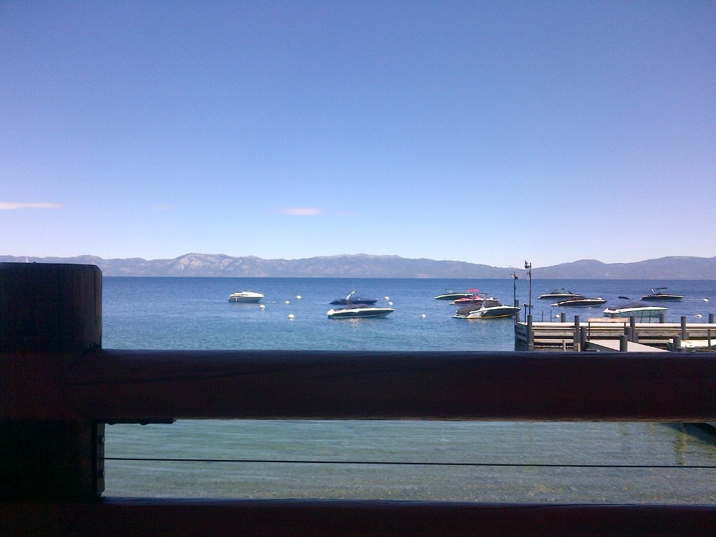 A clear view of the tranquil blue lake is captured, with several boats bobbing gently on the water's surface. The distant mountains provide a scenic backdrop beneath a clear sky, framing the serene aquatic landscape.