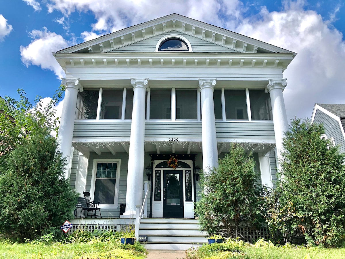 A stately residential house with prominent white columns supports a spacious porch. The entrance features double doors framed by large windows. Lush green shrubbery surrounds the front yard, complemented by decorative elements such as a seasonal wreath and visible outdoor seating on the porch.