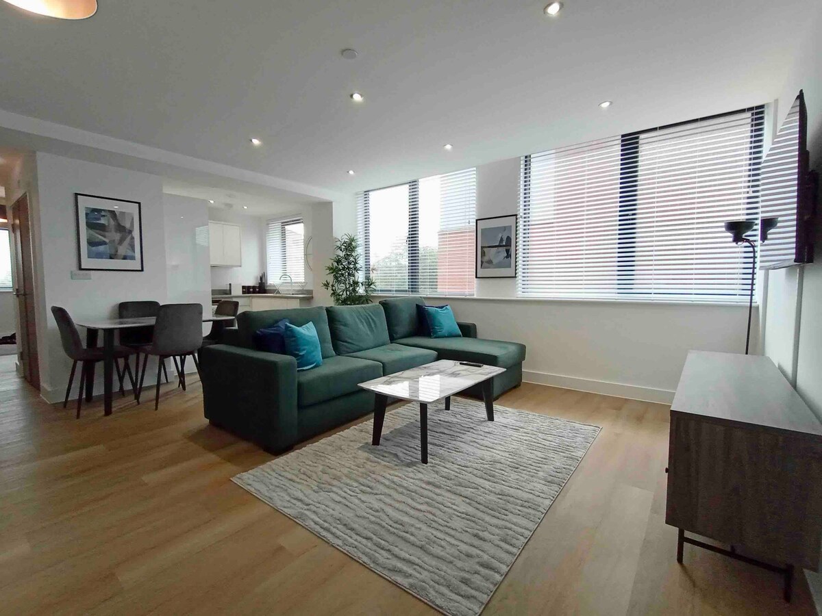 A modern living room features a green sofa with decorative cushions, positioned in front of a wall-mounted TV. Natural light enters through large windows fitted with blinds, illuminating the spacious area. A coffee table rests on a textured rug, while a dining table and chairs are visible in the background.