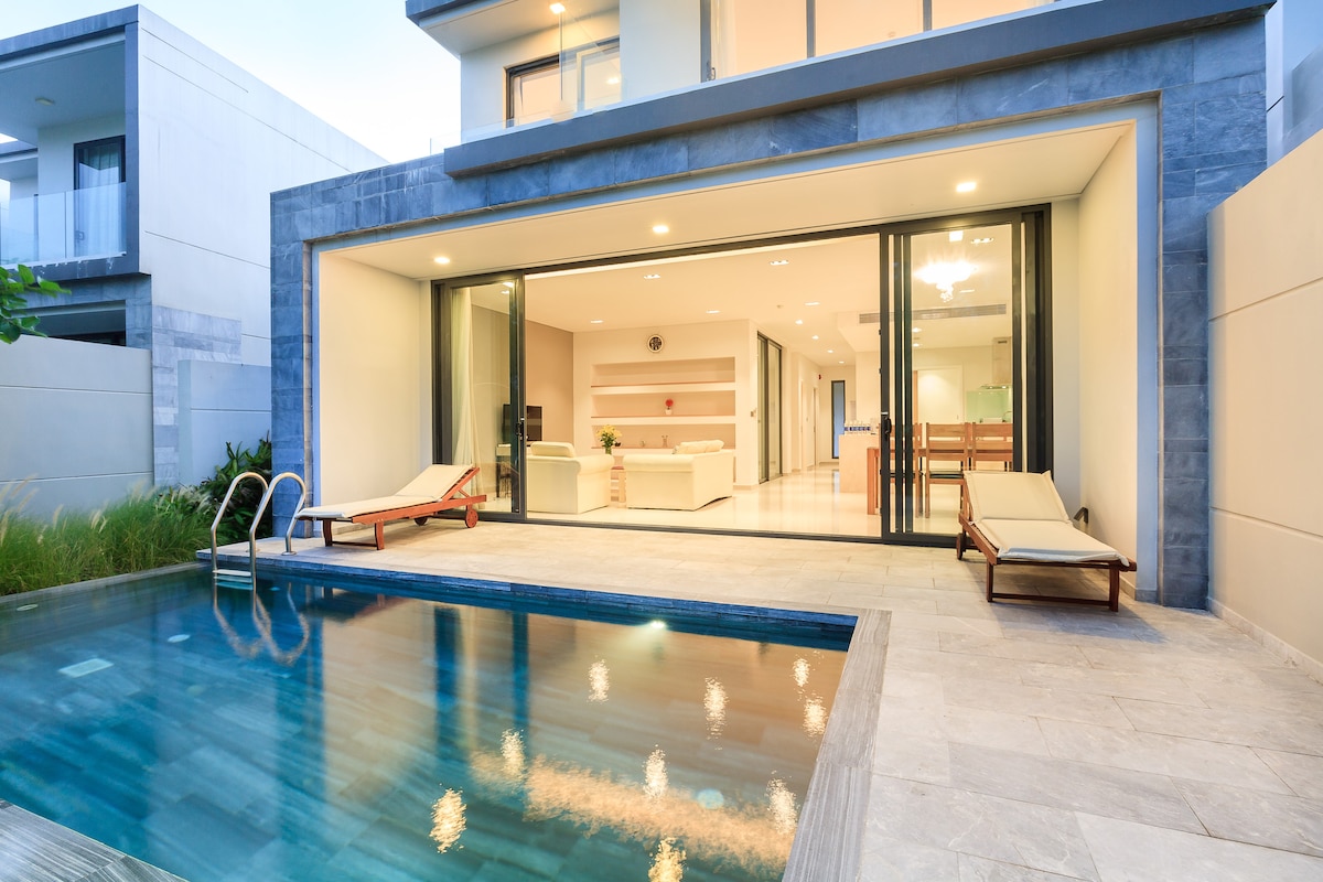 An outdoor area features a private swimming pool with sleek tiles reflecting light. Loungers are positioned beside the water, and expansive glass doors open to a contemporary interior space, creating a seamless transition between indoors and outdoors.