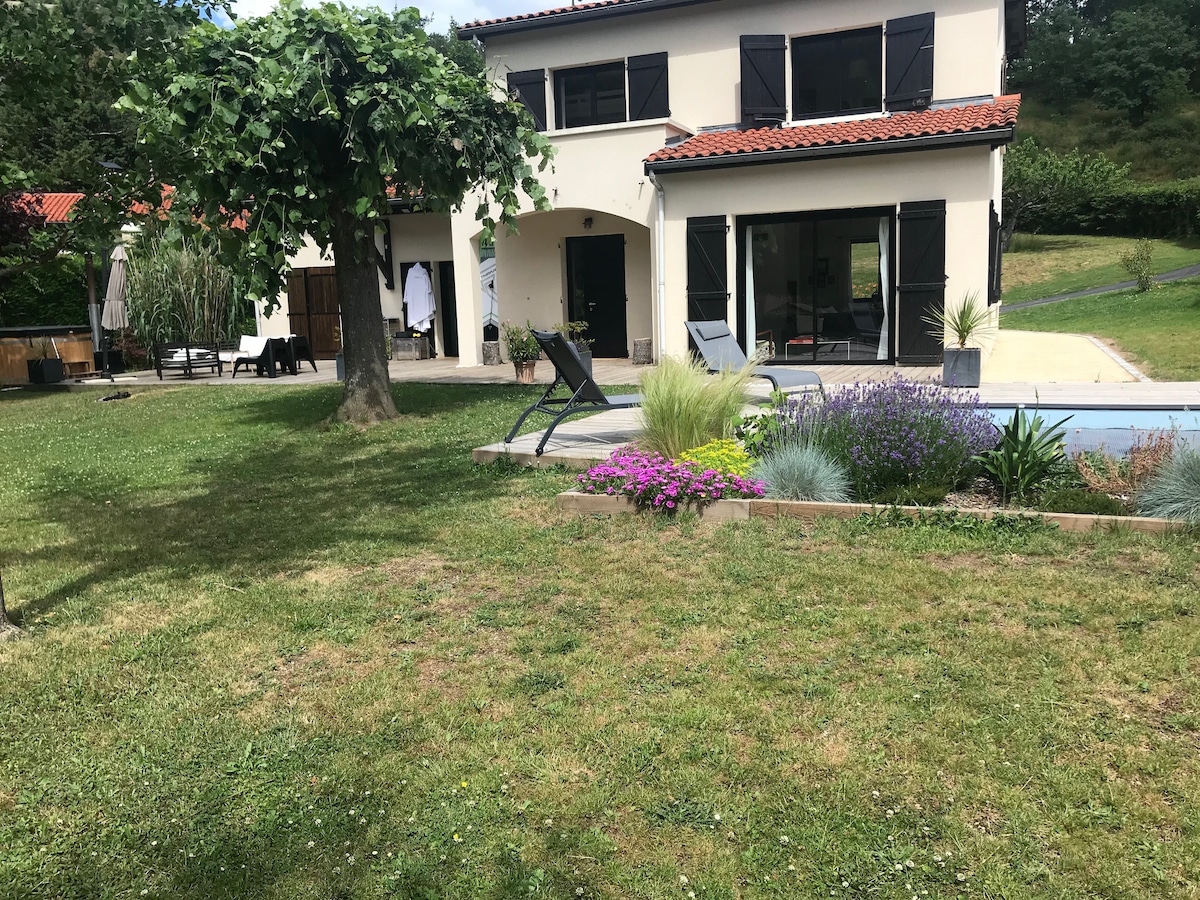A serene outdoor area features a well-maintained garden with colorful flowers and greenery. A two-story house is visible, showcasing large windows and a covered porch. Lounge chairs are positioned near a clear pool, surrounded by lush landscaping and shaded by a tree.