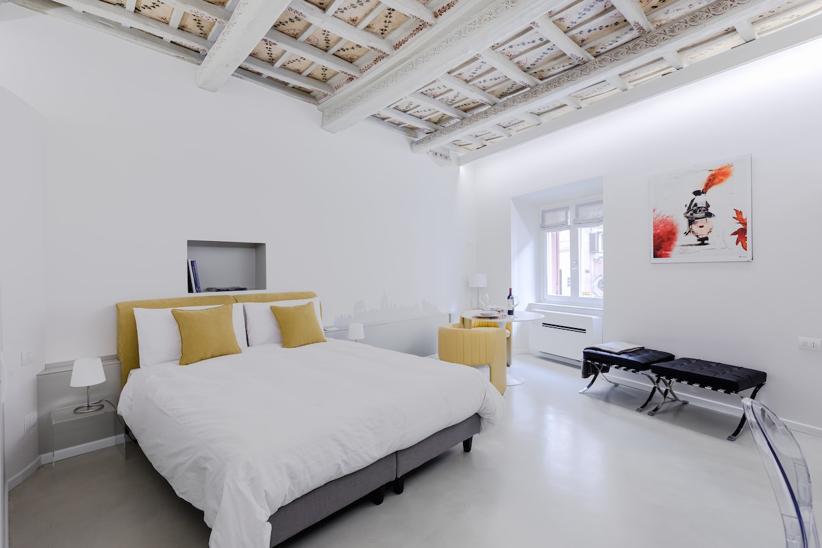A spacious bedroom features a double bed with light bedding and yellow accent pillows. Elegant exposed beams adorn the ceiling, while natural light enters through a nearby window. Two black benches are positioned at the foot of the bed, offering additional seating.