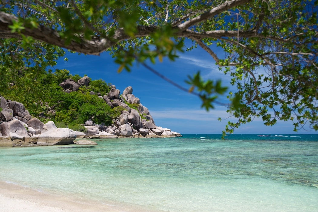 A serene beach scene features clear turquoise waters gently lapping against soft white sand. Lush greenery and rugged rocks create a picturesque backdrop, while the bright blue sky and a few scattered clouds enhance the tranquil atmosphere of this coastal haven.