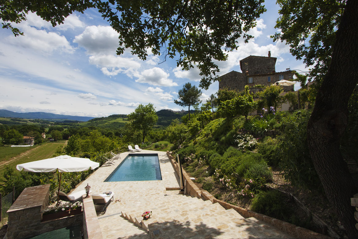 A serene outdoor area features a swimming pool surrounded by lush greenery and scenic views of the rolling hills. A stone pathway leads from the pool to the historical mansion perched above, with a shaded umbrella inviting relaxation.
