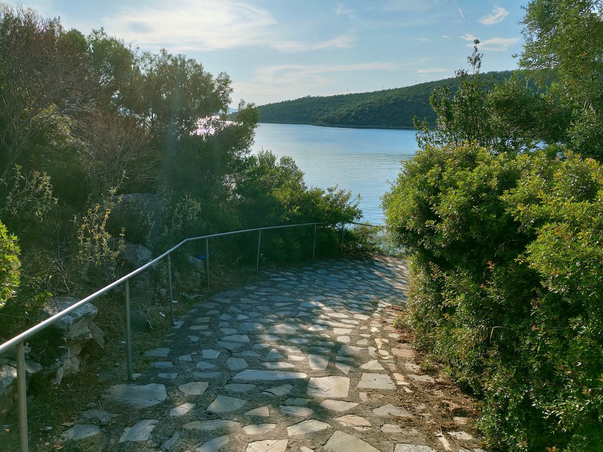 A stone pathway winds gently towards a serene body of water, framed by lush greenery on both sides. Sunlight glistens on the water's surface, and the pathway's natural materials blend harmoniously with the surrounding landscape.