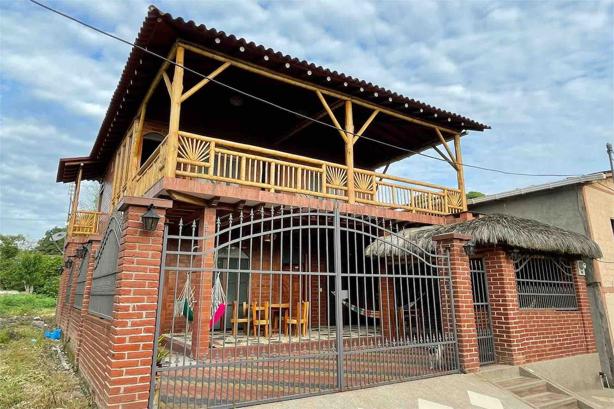 A two-story house is depicted, featuring a wooden balcony with ample space for seating. The facade is primarily brick, complemented by a gated entrance and a thatched roof over the lower area. The surrounding green landscape adds a natural backdrop.