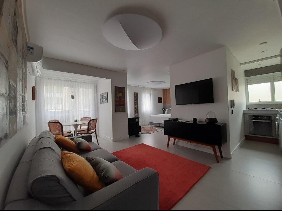 A modern living area is depicted, featuring a comfortable gray sofa adorned with colorful cushions. A vibrant red area rug adds warmth to the space. A dining table for four is visible in the background, while a wall-mounted TV and kitchen area are seen on the right.