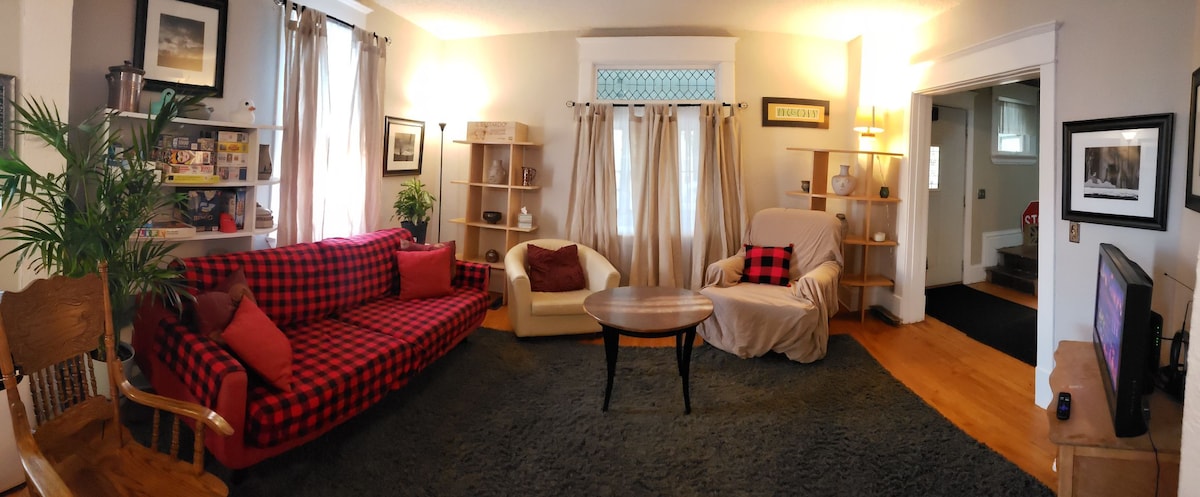 A spacious living area is furnished with a red and black checkered sofa and a cream-colored armchair. A round table sits at the center, surrounded by soft carpet. Natural light filters through window treatments, and wooden shelving displays various decorative items.