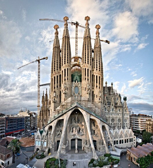 The Sagrada Familia is prominently displayed with its intricate stone carvings and towering spires under construction. Surrounding buildings and palm trees can be seen at ground level, along with cranes indicating ongoing work. The sky is a mix of clouds and blue hues.