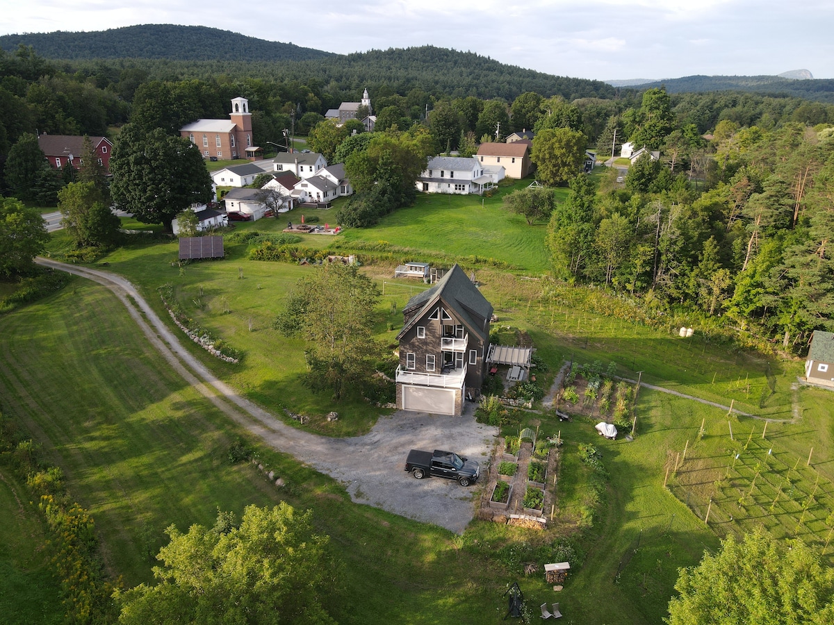An aerial view captures the property with its charming house surrounded by lush greenery and well-maintained gardens. The nearby buildings blend into a peaceful rural setting, complemented by hills in the background, creating a serene environment.