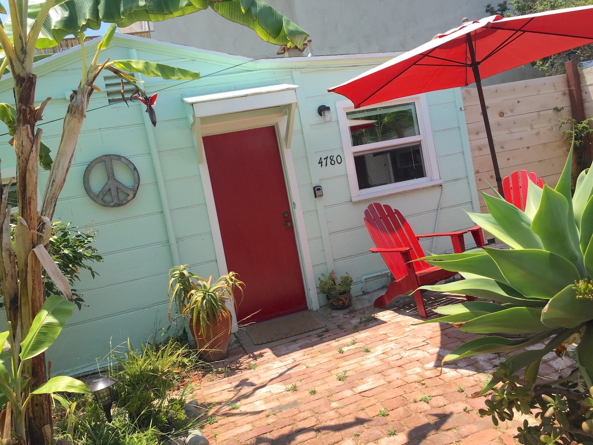 The exterior of a charming tiny home is displayed, featuring a bright red door and vibrant red outdoor chairs. Lush greenery surrounds the patio area, which is paved with brick. A colorful umbrella offers shade, enhancing the inviting outdoor space.