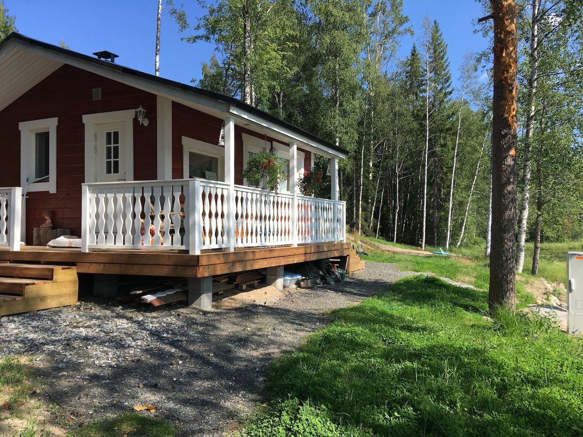 A renovated cabin is set amidst greenery, featuring a large wooden deck with white railings and potted plants. Surrounded by trees, the cabin showcases a red exterior and is designed to blend with the natural landscape.