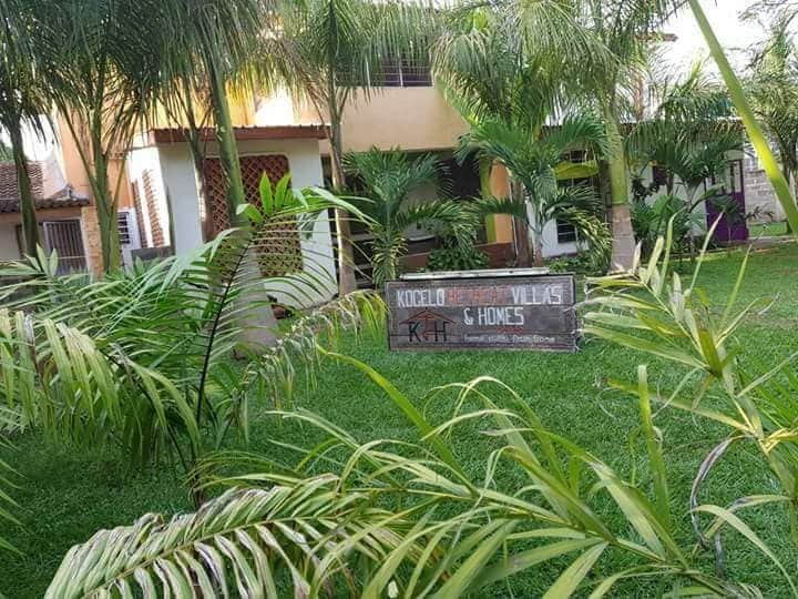 A welcoming entrance showcases the Kogelo Villas sign amidst lush greenery. Towering palm trees and well-maintained grass surround the area, offering a serene atmosphere. The building features a blended architectural style with visible windows and a porch, enhancing the homely vibe of the surroundings.
