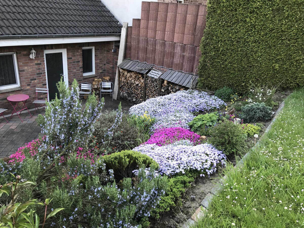 A vibrant garden filled with a variety of colorful flowers in shades of purple, pink, and blue, complemented by neatly stacked firewood against a backdrop of greenery. A small seating area is visible with two chairs and a round table, inviting outdoor relaxation.