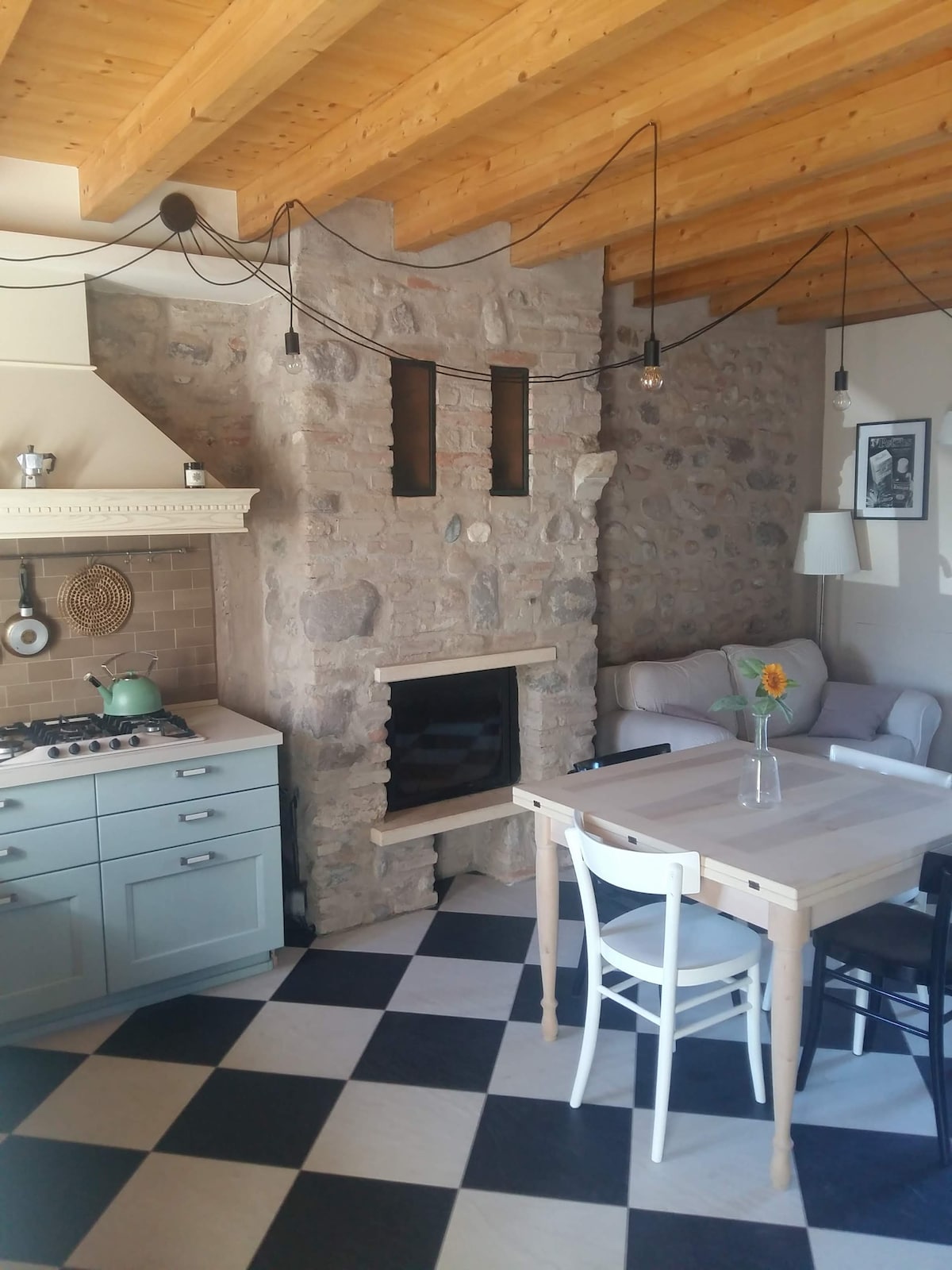 A cozy kitchen and living area is displayed, featuring a rustic stone fireplace. The space contains a light-colored dining table and white chairs. A small green kettle rests on the stove in the kitchen, and a sunflower sits casually on the table, enhancing the inviting ambiance.