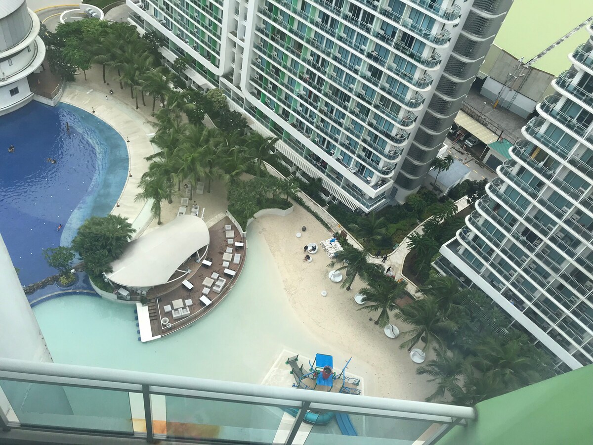 An aerial view captures a resort-style pool area surrounded by palm trees and lounge chairs. A sandy beach area is visible, alongside a shaded structure offering seating. A vibrant blue water slide is situated nearby, contributing to the appealing outdoor space.