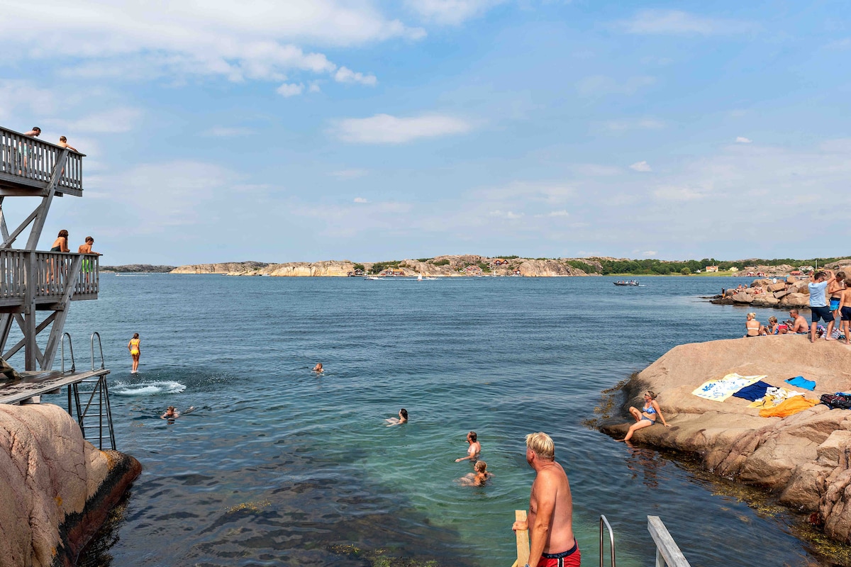 A tranquil bathing area is depicted, featuring rocky outcrops and calm blue waters. Several individuals are seen swimming and enjoying the sun. A wooden platform is visible, allowing easy access to the water, while towels are spread across the rocks nearby.