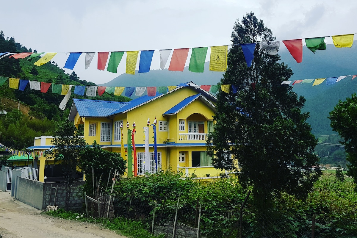 A vibrant yellow house is set against a backdrop of green hills and cloudy skies. Colorful prayer flags are strung across the image, adding a festive touch. Lush greenery surrounds the property, emphasizing its natural setting.