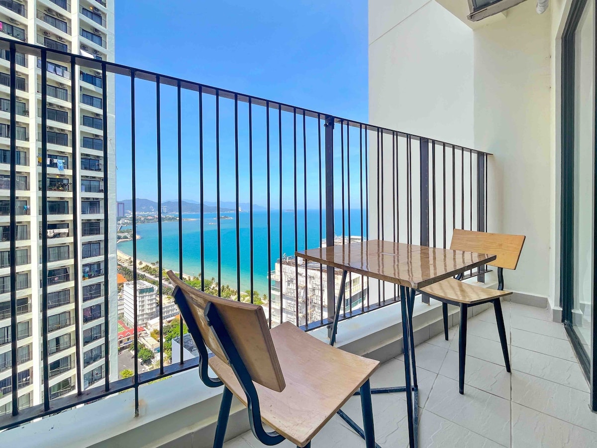 A balcony offers a view of the coastline, with two wooden chairs and a table positioned against a railing. The turquoise sea meets the horizon under a clear blue sky, providing a spacious and airy setting.