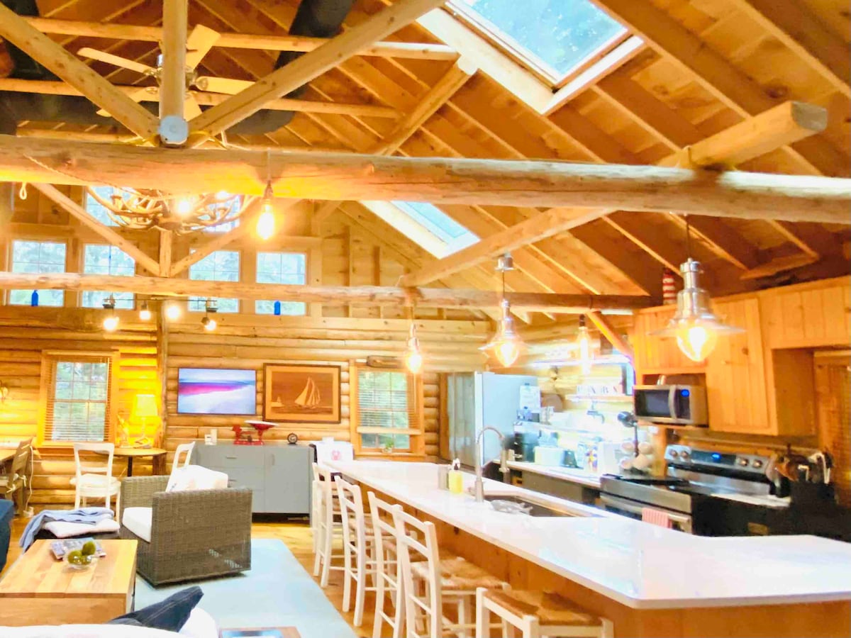 The cabin's interior features wooden beams and a high ceiling, promoting an open atmosphere. Skylights allow natural light to illuminate the space. A modern kitchen is equipped with stainless steel appliances and a large island, while a cozy seating area invites relaxation.