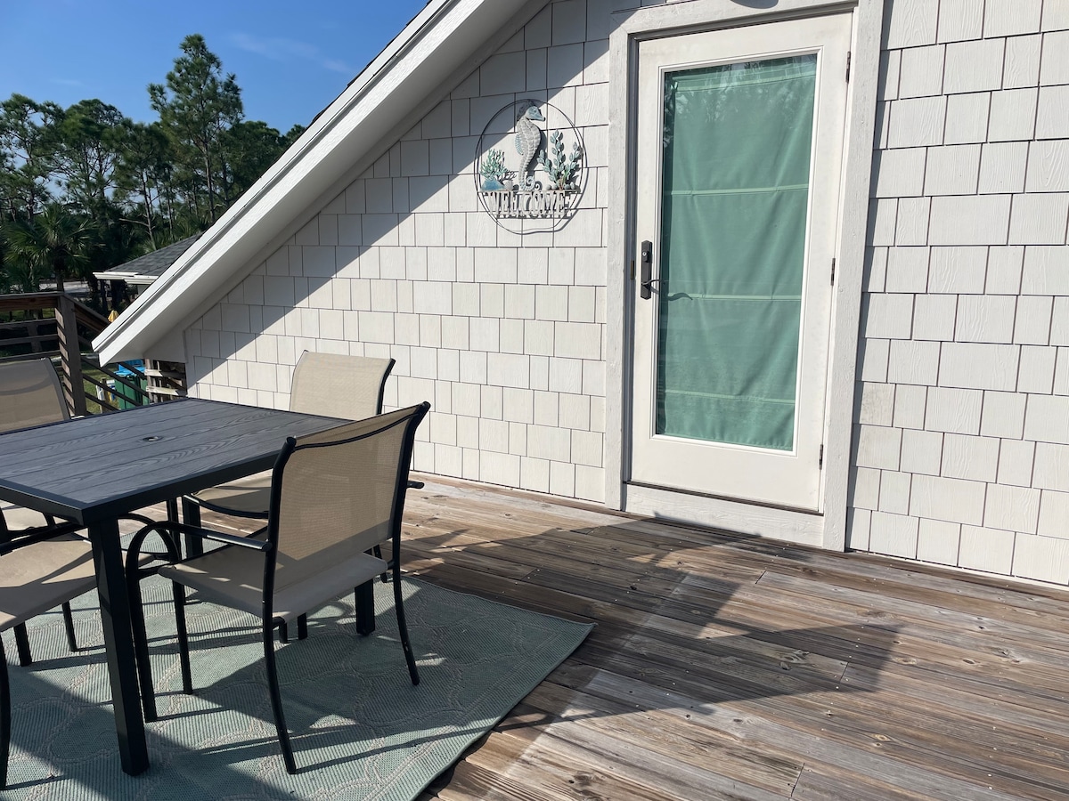 A private balcony features a wooden deck and a table with four chairs, providing a comfortable setting for outdoor relaxation. Sliding glass doors lead into the loft, enhancing access to natural light. The area is surrounded by greenery, offering a serene atmosphere.