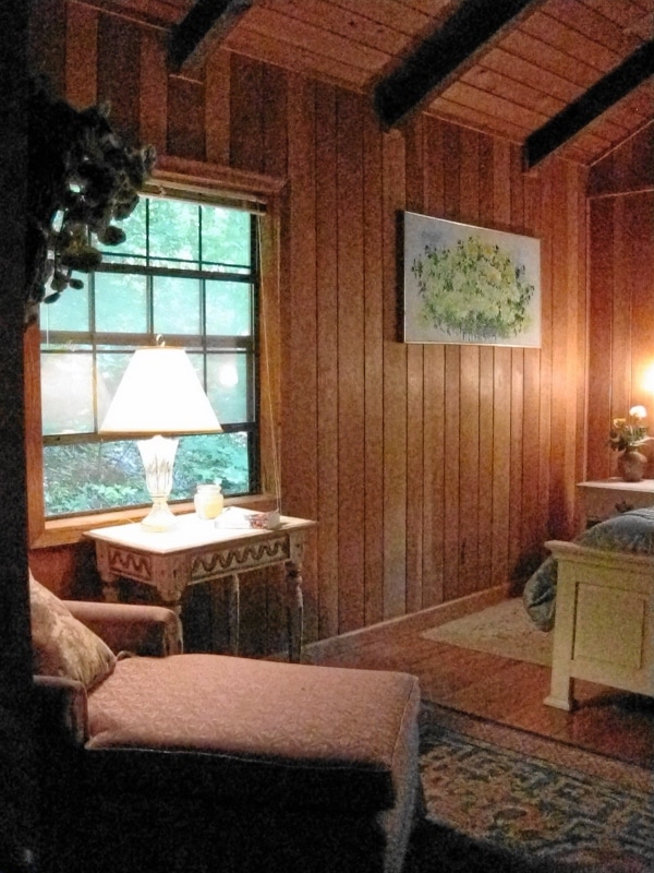 A cozy corner of a wooden room is highlighted by a large window allowing natural light to filter in. A comfortable armchair sits beside a decorative side table adorned with a lamp. A piece of artwork is displayed on the wall, complemented by a soft area rug.