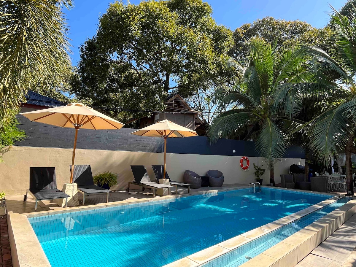 The image displays a swimming pool surrounded by lounge chairs and umbrellas, set against a backdrop of lush greenery. Sunlight reflects off the clear water, enhancing the tranquil atmosphere. The outdoor space is designed for relaxation and enjoyment.