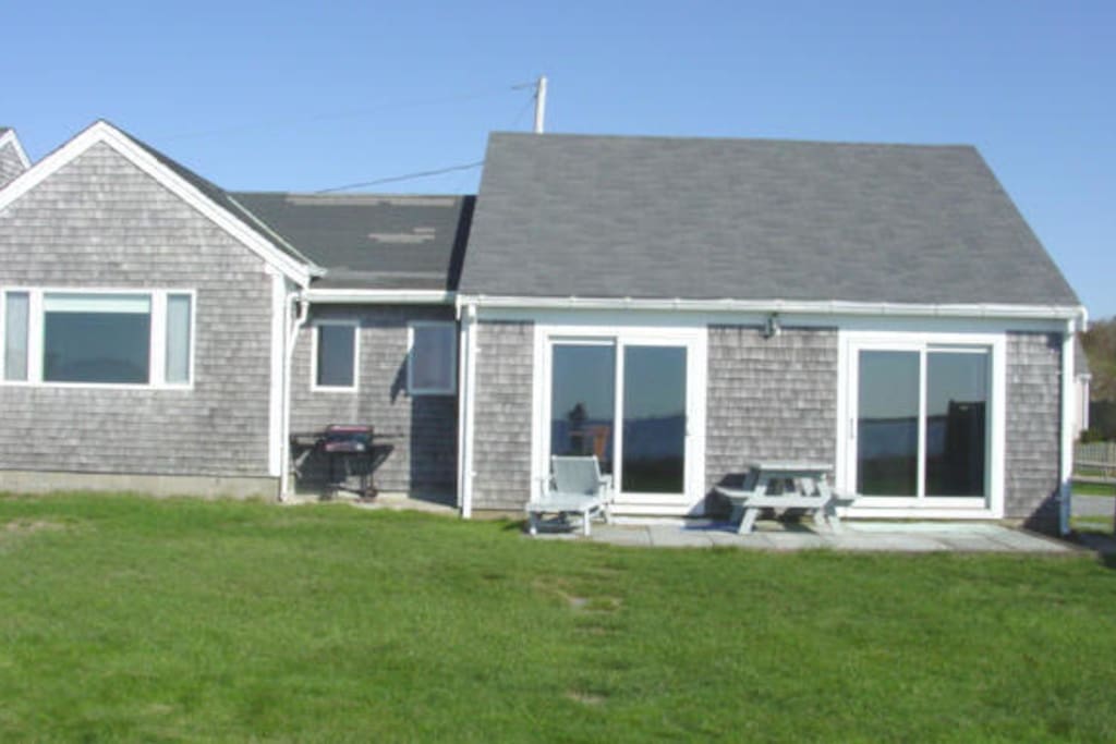 Green lawn overlooks the water. View from the top of the beach steps looking back at the house. That's a patio with picnic table and grill on the left. The bay window on the left is Bedroom 1.