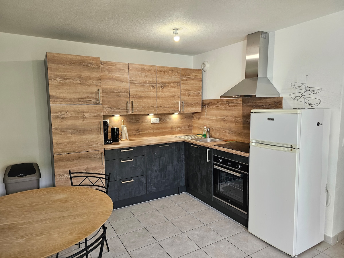 The kitchen features modern cabinetry with a combination of wood and dark finishes. A dining table with metal chairs is positioned to one side, while appliances, including a refrigerator and an oven, are integrated into the layout. Subtle lighting enhances the workspace.