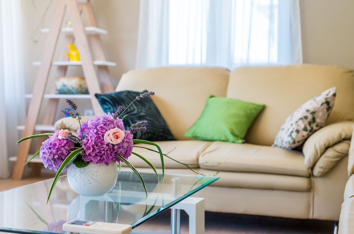A light and airy living area is featured, showcasing a beige sofa adorned with green accent pillows. A glass coffee table is positioned in front, with a vase of vibrant pink and purple flowers adding a splash of color. Soft natural light enters through sheer curtains.