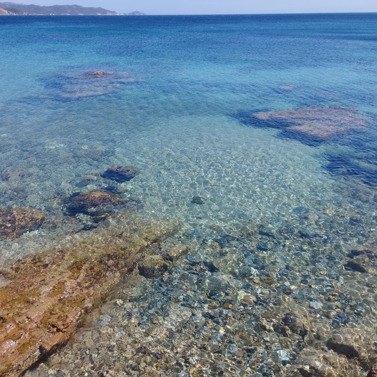 The image displays clear, turquoise waters gently lapping over a rocky seabed. Sunlight filters through the water, creating a shimmering effect on the stones below. Various sizes of smooth rocks and pebbles are visible, enhancing the natural beauty of the coastal environment.