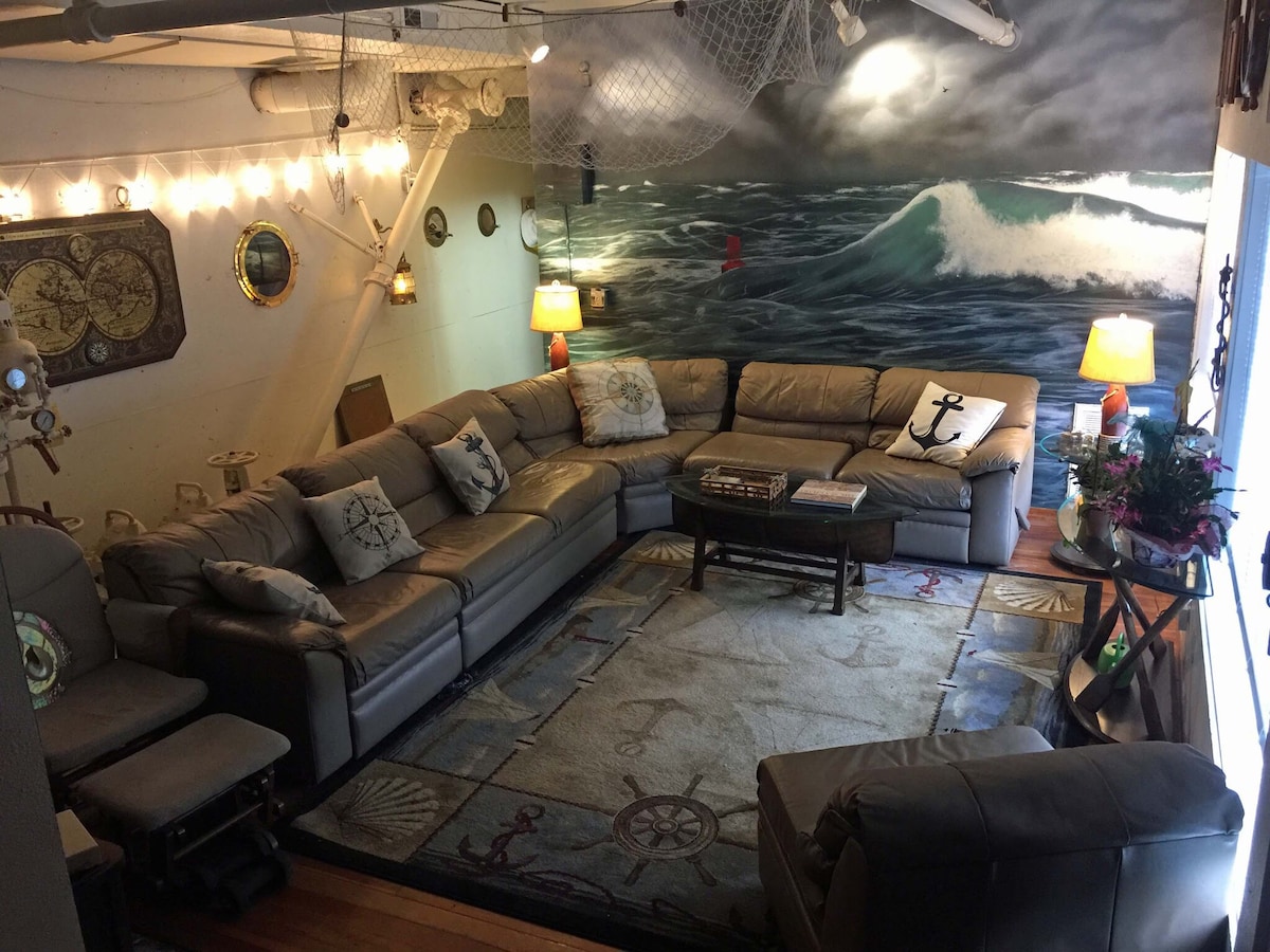The living room area features a spacious sectional sofa and light-toned armchairs, arranged around a decorative area rug. Nautical-themed decor, including pillows and wall art, complements the ocean mural. Soft lighting from table lamps enhances the cozy atmosphere of the space.