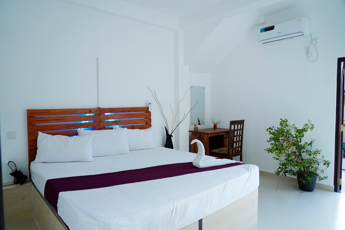 A spacious sleeping area features a large bed with a white duvet and a decorative purple runner. A small wooden desk with a chair is positioned nearby, complemented by a minimalist plant. Natural light enters through the open door, enhancing the bright, neutral decor.