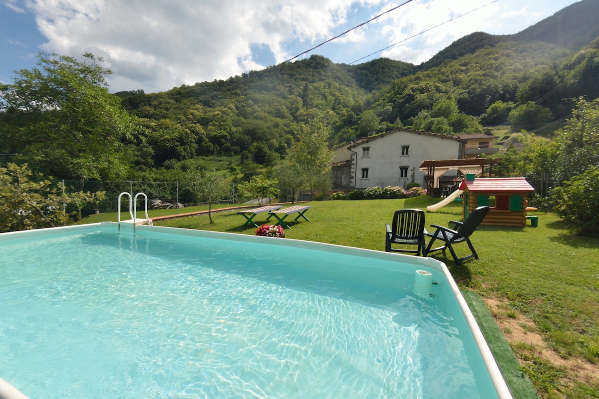 A private swimming pool is visible, surrounded by a lush green garden, with seating options provided by dark chairs. In the background, the holiday home blends harmoniously with the natural landscape of rolling hills, while a play area is designated for children.