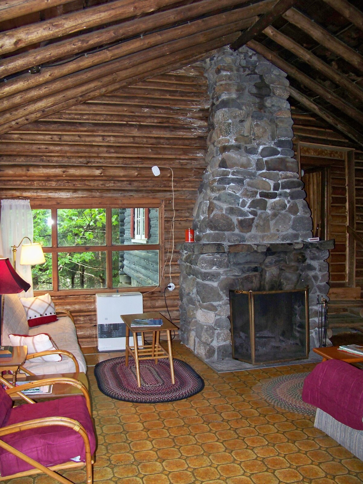 A cozy living area features a stone fireplace as its focal point, surrounded by wooden walls and a sloped ceiling. Two seating options, a couch and a chair, are arranged on a textured rug beside a small table, with natural light filtering through the windows.
