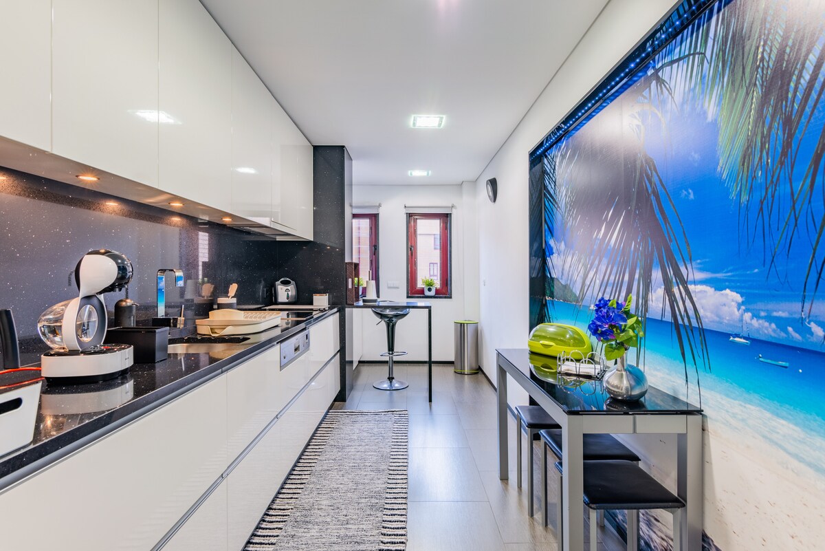 A modern kitchen features sleek white cabinetry and a dark countertop. A striking mural of a beach scene with palm trees creates a vibrant backdrop. A small dining area is furnished with a table and two chairs, complemented by decorative elements like a blue vase.