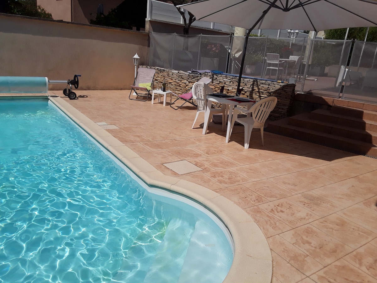 A swimming pool is surrounded by a tiled patio featuring a table and four chairs under a large umbrella. Two lounge chairs are positioned nearby, with a stone wall adding texture to the space. A gentle sparkle reflects off the water's surface.