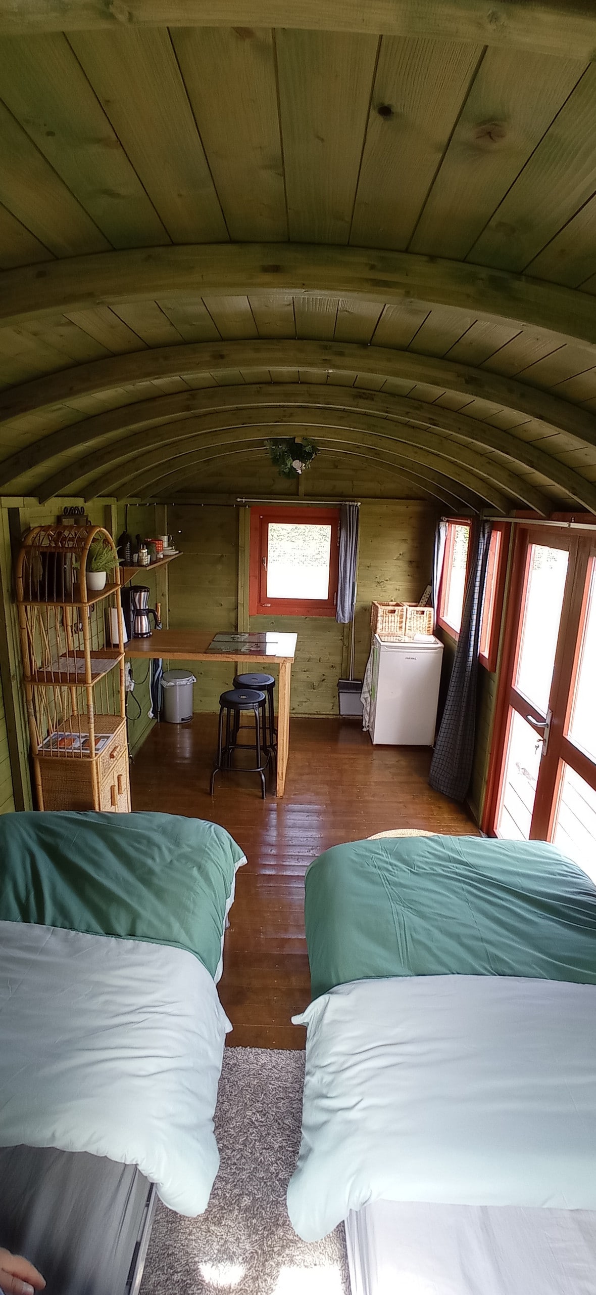 The interior of the tiny house features a wood-paneled design with a curved ceiling. Two separate beds are positioned on the left, dressed in light green linens. A small dining table with a bar stool is visible, along with a refrigerator and a shelf, creating a functional living space.