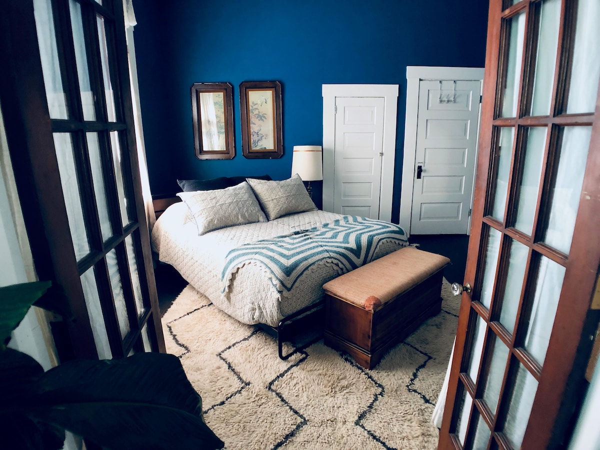 A cozy bedroom is framed by two sets of French doors, featuring a spacious bed dressed with a patterned blanket. The wall is adorned with vintage mirrors. A textured rug covers the floor, while a wooden bench is positioned at the foot of the bed, adding warmth to the space.
