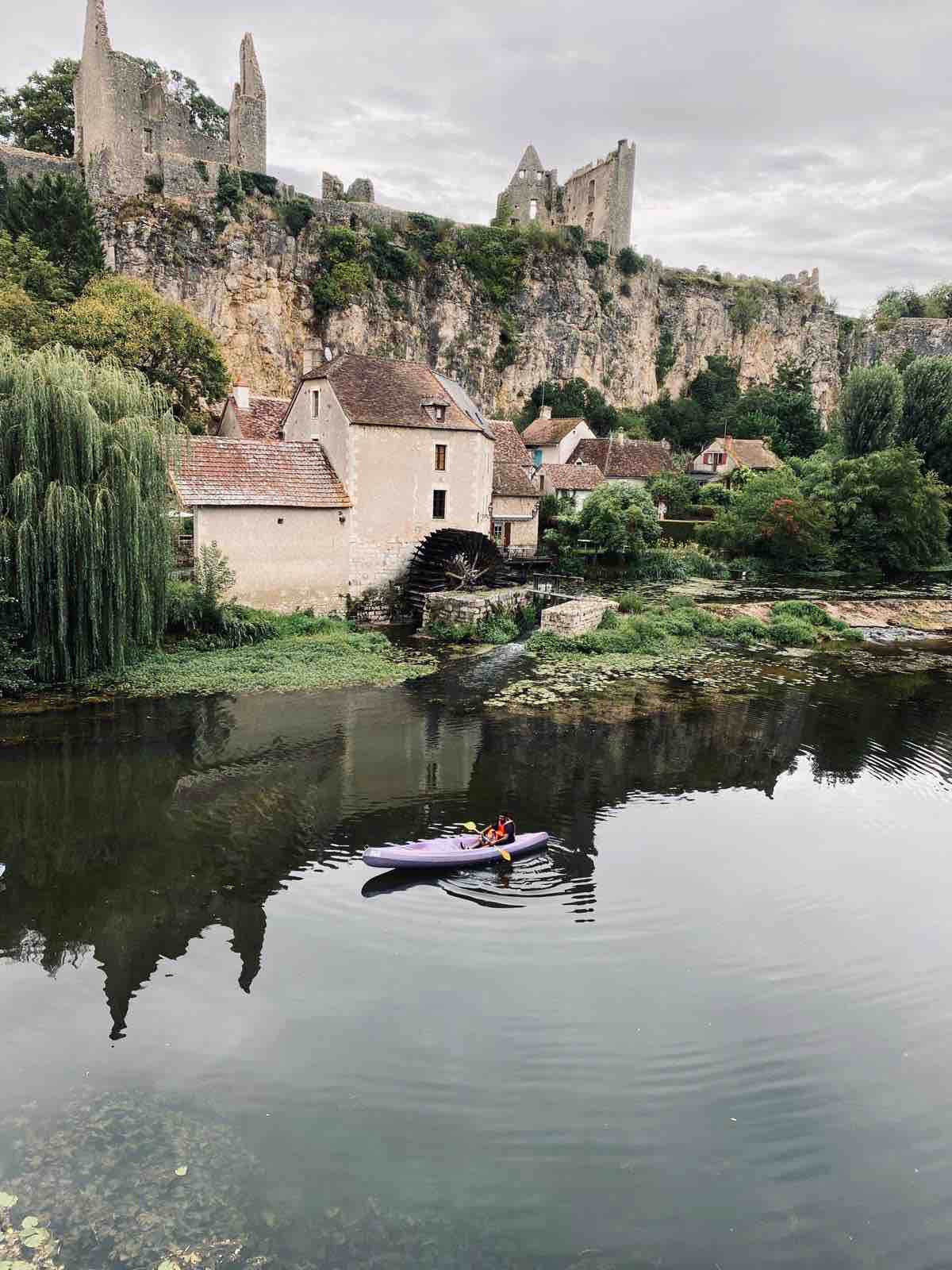 A tranquil river scene is presented, featuring a person in a kayak gliding across the calm water. Lush greenery lines the banks, while an old watermill and charming stone houses are nestled along the riverfront, with medieval ruins perched atop the cliffs in the background.