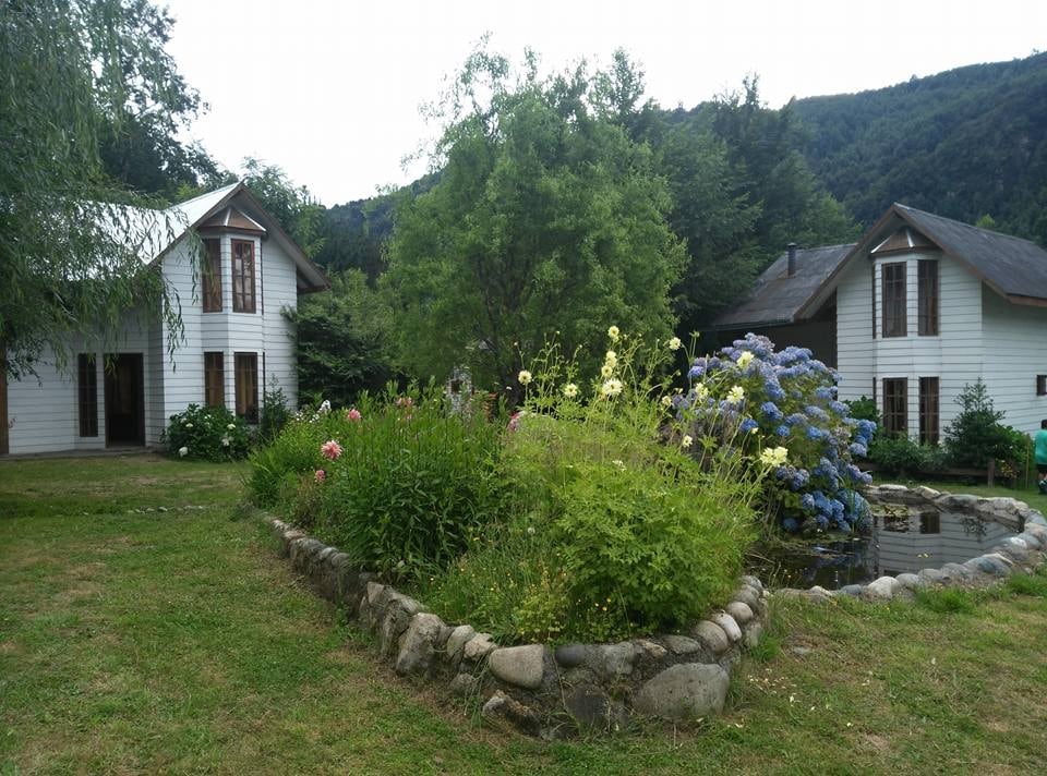 Two charming cabins are set among lush greenery, surrounded by colorful flowering plants. A small pond is visible nearby, enhancing the tranquil atmosphere. The mountainous backdrop adds depth to this serene location, creating a peaceful retreat for relaxation.