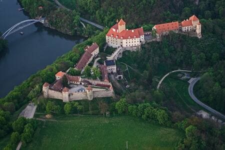 An aerial view captures a historical castle complex surrounded by lush greenery and a winding river. The structures display varied architectural styles, with a patchwork of rooftops colored in red. A road traces along the perimeter, highlighting the serene landscape.