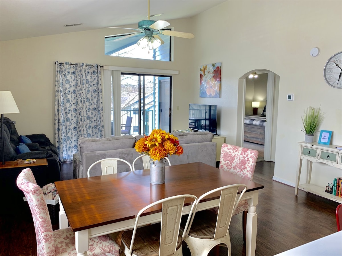 An open living area is presented, featuring a wooden dining table surrounded by six chairs, adorned with a vase of bright flowers. A comfortable gray sofa and a ceiling fan are visible, along with large windows allowing natural light to fill the space.