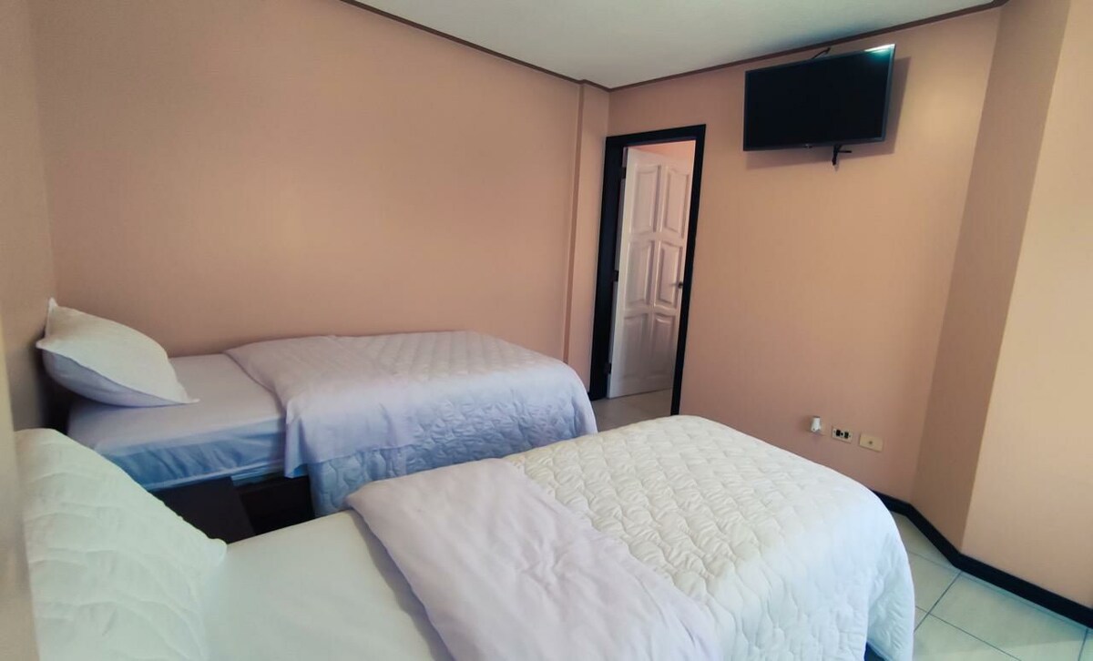A room featuring two comfortable beds with light bedding, positioned parallel to each other. A flat-screen television is mounted on the wall opposite, and the walls are painted in a soft peach color, contributing to a calm and inviting environment.